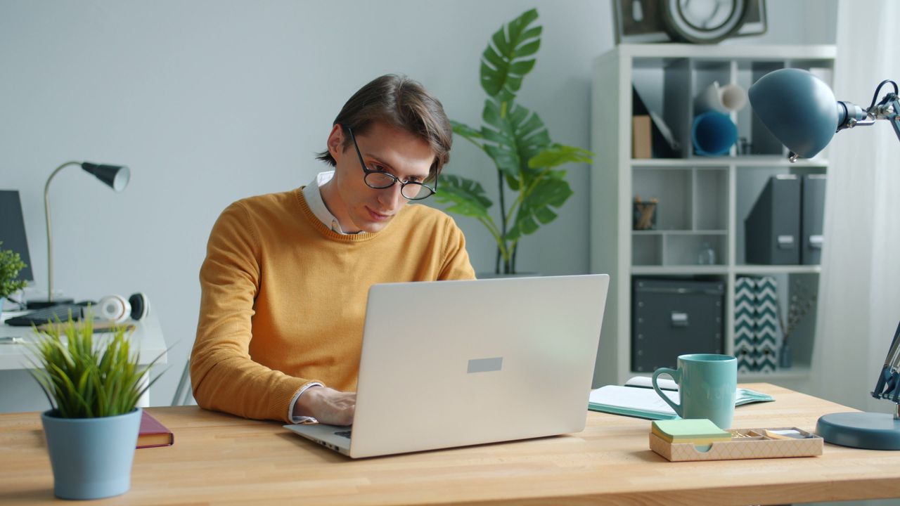 A solo professional working at a laptop on a wooden desk in a bright home office.
